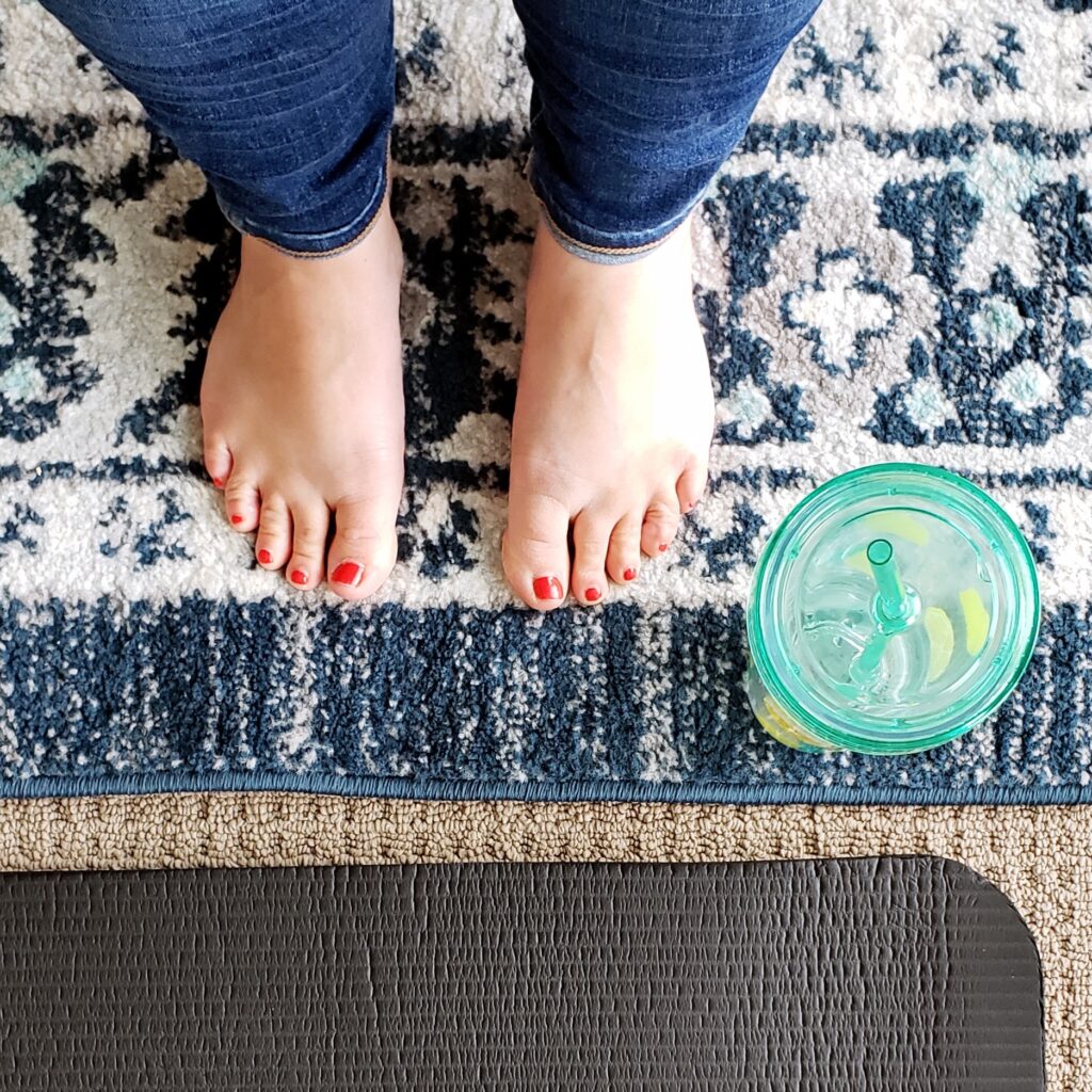 A woman's view looking down at her feet as she stands in front of a black yoga mat and a mint green water bottle.